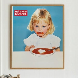 Peut inclure: Une affiche encadrée représentant un enfant blond mangeant de la soupe, avec le texte "eat more borscht" dans un encadré rouge. L'affiche est accrochée à un mur blanc au-dessus d'un comptoir blanc avec deux tabourets tressés.