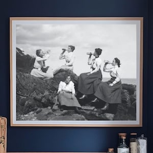 May include: A black and white photograph of five women standing on a rocky beach, all wearing dresses and holding drinks. The photo is framed and hanging on a dark blue wall in a kitchen.