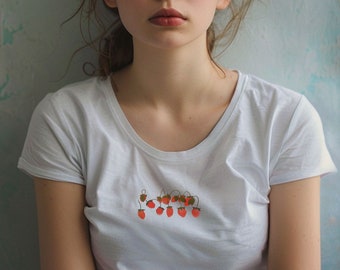 Könnte beinhalten: Eine junge Person mit langen braunen Haaren trägt ein weißes T-Shirt mit einem kleinen gestickten Design aus roten Erdbeeren.