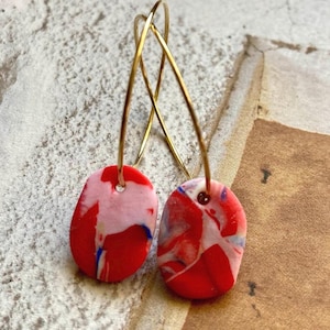 Puede incluir: Un par de pendientes de aro dorados con colgantes de arcilla marmoleada en rojo, blanco y azul. Los colgantes tienen forma ovalada y un diseño único y abstracto.