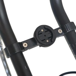 Op de afbeelding: Zwarte fietscomputerhouder bevestigd aan het stuur. De houder is gemaakt van zwart kunststof en heeft een cirkelvormige basis met een reeks sleuven en gaten voor het bevestigen van een fietscomputer. De houder wordt met twee klemmen aan het stuur bevestigd.