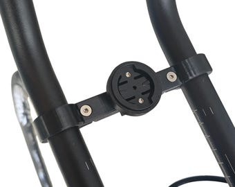 May include: Black bicycle computer mount attached to the handlebars. The mount is made of black plastic and has a circular base with a series of slots and holes for securing a cycling computer. The mount is secured to the handlebars with two clamps.