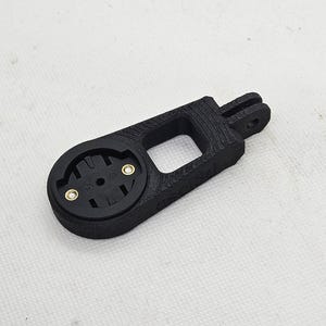 May include: Black 3D-printed bicycle computer mount. The mount has a circular base with two gold screw inserts, a rectangular opening, and a fork-like attachment. It is designed to hold a bicycle computer.