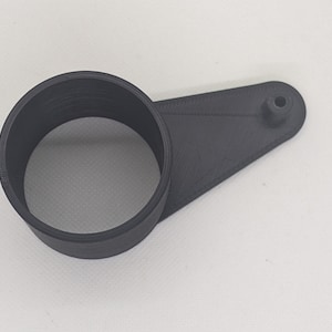 Può includere: Un oggetto nero stampato in 3D con un anello circolare collegato a un pezzo triangolare piatto con una piccola sporgenza cilindrica. L'oggetto sembra essere realizzato in un materiale opaco e strutturato. Il design suggerisce un componente funzionale, possibilmente meccanico.