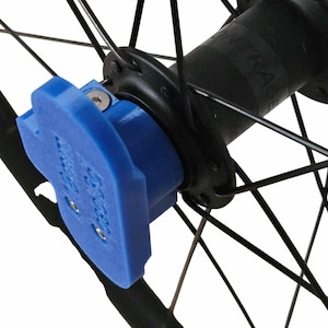 Può includere: Primo piano di una ruota di bicicletta con un componente blu attaccato al mozzo. Il componente ha una forma unica ed è fissato con viti. I raggi della ruota sono neri e anche il mozzo è nero.