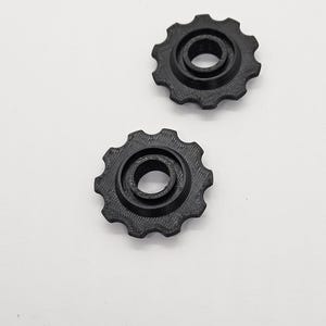 May include: Two black, gear-shaped components with a central hole and toothed outer edges are shown against a white background. The gears appear to be made of a textured material, possibly plastic or a similar substance.