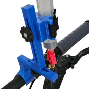 Può includere: Uno strumento blu per lo spurgo dei freni della bicicletta fissato al manubrio della bicicletta. Lo strumento comprende un serbatoio di plastica trasparente, un raccordo rosso e una manopola di regolazione nera. La leva del freno della bicicletta è visibile sullo sfondo.