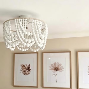 May include: A ceiling light fixture featuring multiple draped strands of matte white wooden beads suspended from a circular frame, creating a tiered, scalloped effect. The fixture is mounted to the ceiling above a bed with pink and white pillows.