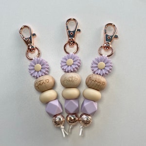 Könnte beinhalten: Drei Schlüsselanhänger mit Holzperlen, lila Blumenanhängern und goldfarbenen Metallschließen. Die Holzperlen sind mit den Worten "Schwester", "Tante" und "Mama" mit einem Herzsymbol graviert.