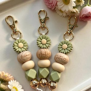 May include: Three keychains featuring gold-tone lobster clasps, sage green silicone flower beads, engraved wooden beads labeled Mama, Aunt, and Sister, cream-colored silicone beads, sage green geometric silicone beads, and gold metallic spheres.