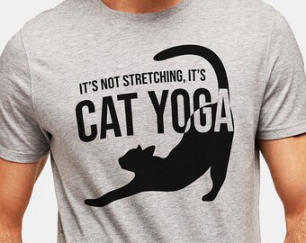 Peut inclure: Un t-shirt gris avec une silhouette noire d'un chat s'étirant en position de yoga. Le texte "It's not stretching, it's cat yoga" est imprimé sur le t-shirt.