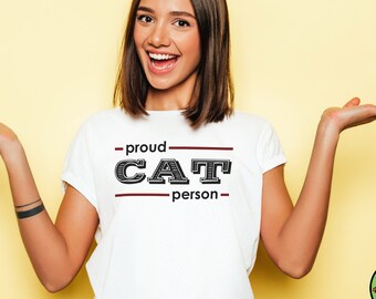 Peut inclure: Un t-shirt blanc avec le texte "proud cat person" en lettres noires et rouges. Le t-shirt est porté par une personne aux cheveux bruns et souriante. L'arrière-plan est jaune clair.