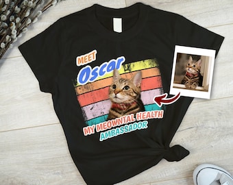 Pode incluir: Camiseta preta com um design retro de arco-íris e o texto "Meet Oscar, My Meowntial Health Ambassador". Uma foto de um gato tigrado está na camiseta.