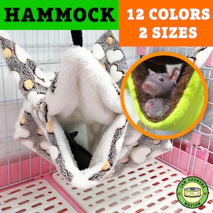 May include: A grey and white plush hammock for small pets, hanging inside a pink cage. The hammock has a heart pattern and is designed to be cosy and comfortable. The text "HAMMOCK 12 COLORS 2 SIZES" is visible in the image.