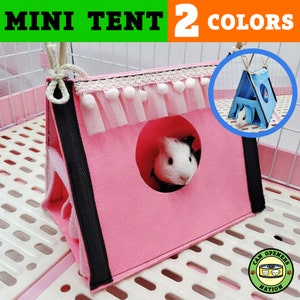 May include: A pink and blue mini tent for small pets, with a white lace trim and pom poms. The tent has a round opening and is shown with a guinea pig inside. The text "MINI TENT 2 COLORS" is visible at the top of the image.