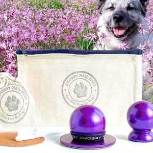 May include: A set of two purple dog nail files in a small canvas bag with the text "KNOBBY NAIL FILES Easy Nail Care for Dogs!" printed on it. The files are on a white surface with a cork coaster and a white nail file.
