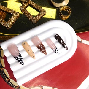 May include: A collection of five artificial nails with various designs. The nails feature colors like nude, brown, and black, with accents of rhinestones and polka dots. The nails are displayed on a white, rectangular tray, with gold jewelry in the background.