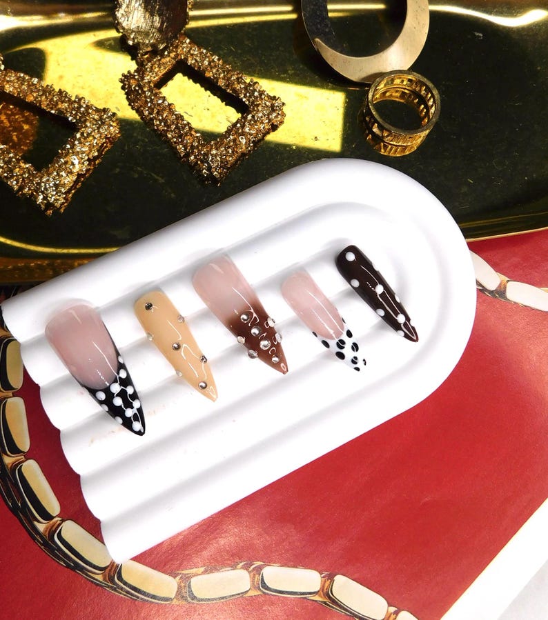May include: A collection of five artificial nails with various designs. The nails feature colors like nude, brown, and black, with accents of rhinestones and polka dots. The nails are displayed on a white, rectangular tray, with gold jewelry in the background.