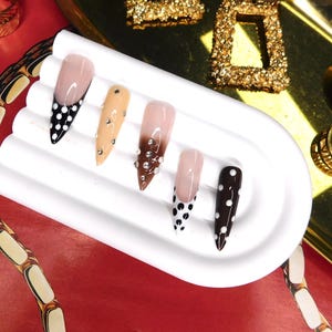 May include: A collection of long, pointed artificial nails in various designs. Some nails feature a nude base with black polka dots, while others have embellishments or a gradient effect. The nails are displayed on a white, curved platform, with gold jewelry in the background.