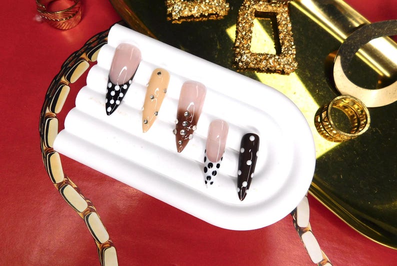 May include: A collection of long, pointed artificial nails in various designs. Some nails feature a nude base with black polka dots, while others have embellishments or a gradient effect. The nails are displayed on a white, curved platform, with gold jewelry in the background.