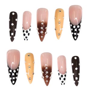 May include: A set of ten artificial nails in various designs. Some nails feature a brown base with white polka dots, while others have a nude base with a black polka dot design. Several nails have a nude to brown gradient with rhinestones.