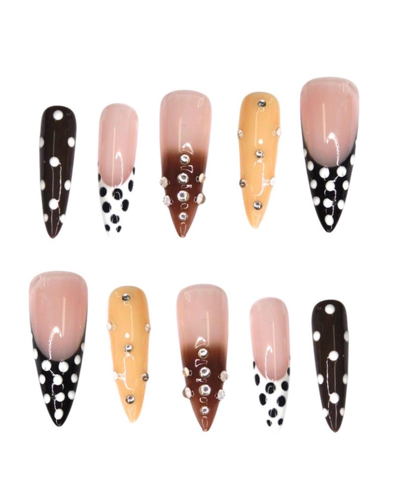 May include: A set of ten artificial nails in various designs. Some nails feature a brown base with white polka dots, while others have a nude base with a black polka dot design. Several nails have a nude to brown gradient with rhinestones.