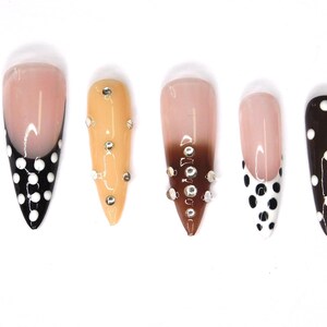 May include: A set of five artificial nails in various designs. One nail features a black base with white polka dots, while another has a French tip with black polka dots. Other nails have a nude base with rhinestones or a brown ombre.