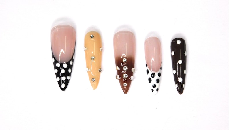 May include: A set of five artificial nails in various designs. One nail features a black base with white polka dots, while another has a French tip with black polka dots. Other nails have a nude base with rhinestones or a brown ombre.