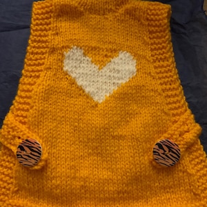 Pode incluir: Um suéter de malha amarelo mostarda com um desenho de coração branco na frente. O suéter tem gola alta e botões decorativos com um padrão de zebra nas mangas. A peça de roupa é feita à mão.