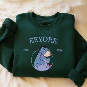 May include: A dark green sweatshirt with the word "Eeyore" embroidered in light blue, above an image of Eeyore. The sweatshirt also features "EST. 1926". The sleeves are cuffed. The sweatshirt is folded on a cream-colored surface.