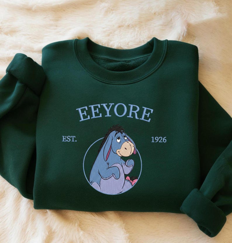 May include: A dark green sweatshirt with the word "Eeyore" embroidered in light blue, above an image of Eeyore. The sweatshirt also features "EST. 1926". The sleeves are cuffed. The sweatshirt is folded on a cream-colored surface.