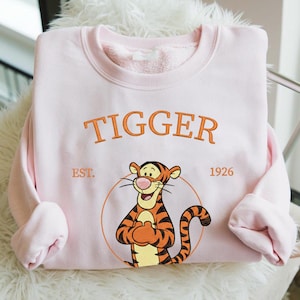 May include: A light pink sweatshirt featuring the word "TIGGER" in orange embroidery above a cartoon tiger character. The tiger is inside a circle, with "EST. 1926" below. The sweatshirt has a crew neck and long sleeves.
