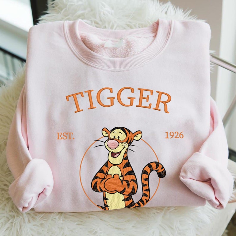 May include: A light pink sweatshirt featuring the word "TIGGER" in orange embroidery above a cartoon tiger character. The tiger is inside a circle, with "EST. 1926" below. The sweatshirt has a crew neck and long sleeves.