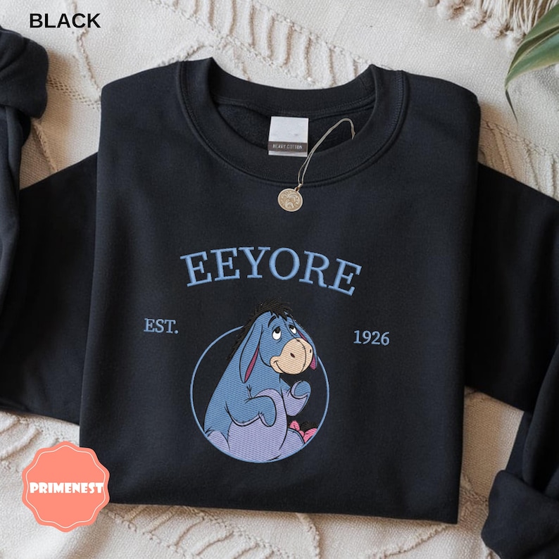 May include: Black sweatshirt featuring the word "Eeyore" in light blue, with "EST. 1926" and a circular graphic of Eeyore. The sweatshirt is made of heavy cotton.