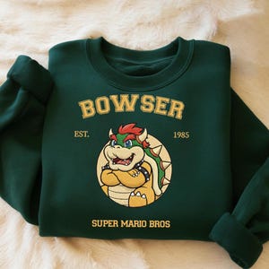 May include: Dark green sweatshirt with a Bowser graphic from Super Mario Bros. The design includes the word "BOWSER" above an image of the character, with "EST. 1985" and "SUPER MARIO BROS" below. The sweatshirt has a crew neck and long sleeves.