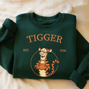 May include: A dark green sweatshirt featuring the word "TIGGER" in orange lettering above an illustration of Tigger from Winnie the Pooh. The sweatshirt also includes the text "EST. 1926" and a circular design around the character.