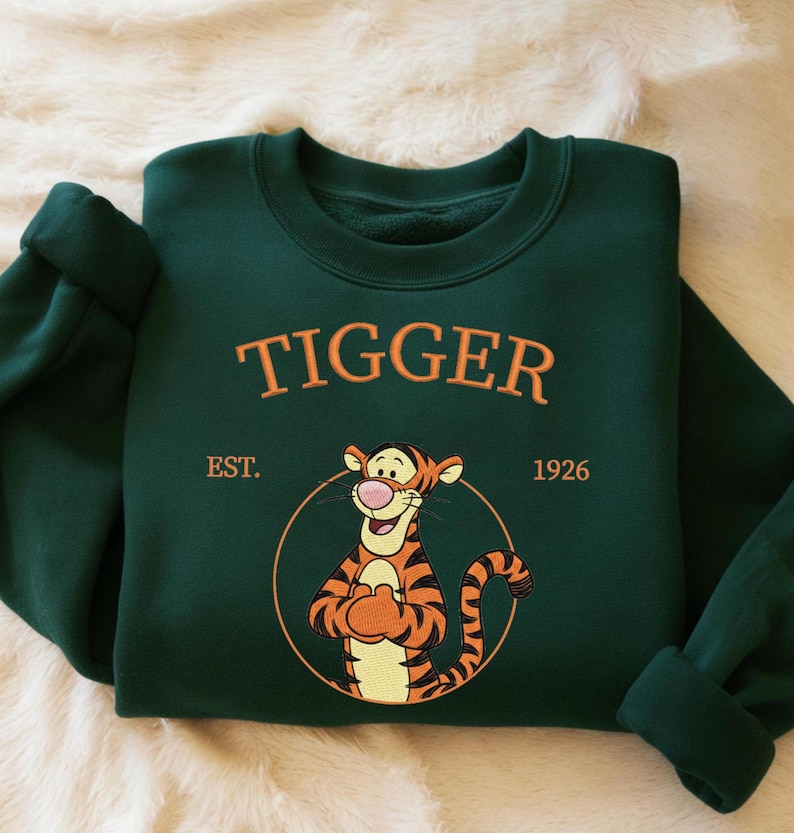 May include: A dark green sweatshirt featuring the word "TIGGER" in orange lettering above an illustration of Tigger from Winnie the Pooh. The sweatshirt also includes the text "EST. 1926" and a circular design around the character.