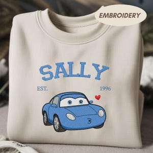 Könnte beinhalten: Cremefarbenes Sweatshirt mit dem Namen "SALLY" in Blau gestickt, zusammen mit einem blauen Cartoon-Auto und einem roten Herz. Der Text "EST. 1996" ist ebenfalls in Blau gestickt.