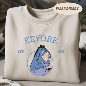 May include: A cream-coloured sweatshirt with an embroidered Eeyore design. The design includes the name "Eeyore" in blue, the year "1926", and the word "Embroidery". The sweatshirt is folded.