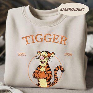 May include: Cream-colored sweatshirt featuring an embroidered image of Tigger from Winnie the Pooh. The word "TIGGER" is embroidered in orange above the character, with "EST. 1926" below. The design includes a circular outline around the character.