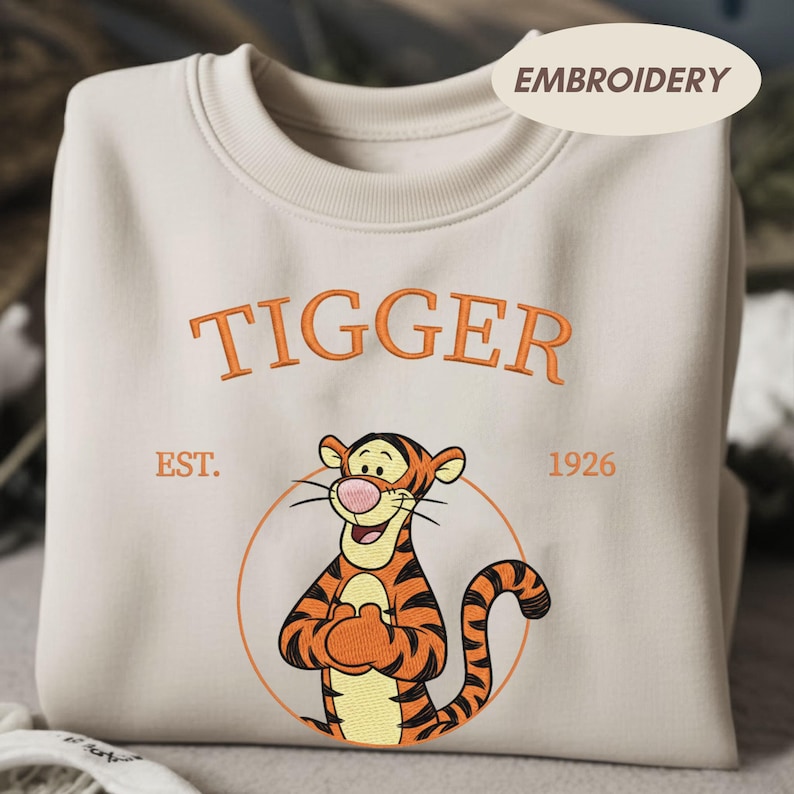 May include: Cream-colored sweatshirt featuring an embroidered image of Tigger from Winnie the Pooh. The word "TIGGER" is embroidered in orange above the character, with "EST. 1926" below. The design includes a circular outline around the character.