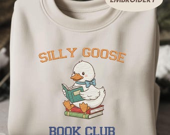 May include: A cream-coloured sweatshirt with the embroidered text "SILLY GOOSE BOOK CLUB." The design features a cartoon duck reading a book while sitting on a stack of books. The duck is wearing a blue bow tie.