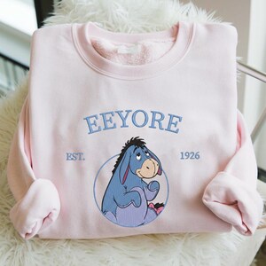 May include: Pale pink sweatshirt with an embroidered Eeyore design. The design includes the name "Eeyore" in blue script above the character, with "EST. 1926" below. The sweatshirt has a crew neck and long sleeves. A classic cartoon character.