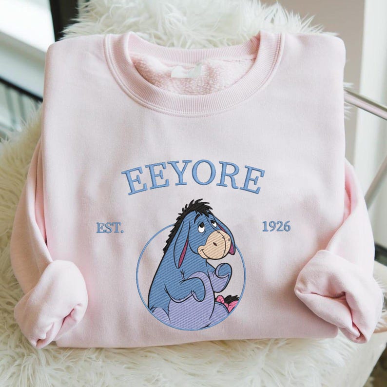 May include: Pale pink sweatshirt with an embroidered Eeyore design. The design includes the name "Eeyore" in blue script above the character, with "EST. 1926" below. The sweatshirt has a crew neck and long sleeves. A classic cartoon character.