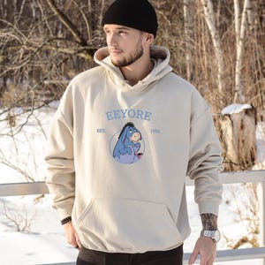 May include: Beige hoodie with a kangaroo pocket and a drawstring hood. The front features a blue and purple cartoon donkey graphic with the text "Eeyore Est. 1926" in light blue. The hoodie is made of a soft material.