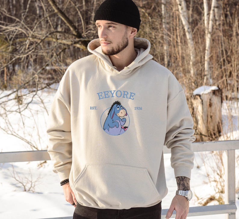 May include: Beige hoodie with a kangaroo pocket and a drawstring hood. The front features a blue and purple cartoon donkey graphic with the text "Eeyore Est. 1926" in light blue. The hoodie is made of a soft material.