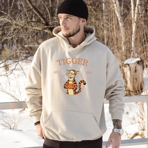 May include: Beige hoodie featuring the word "Tigger" in orange, with an illustration of Tigger from Winnie the Pooh. The hoodie has a kangaroo pocket and a drawstring hood. The text "EST. 1926" is also visible.
