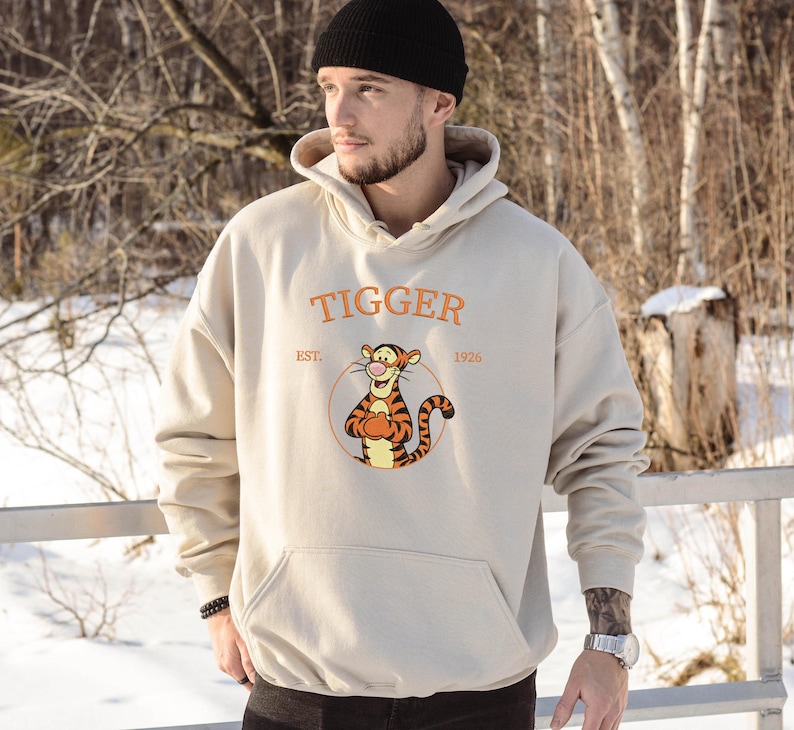 May include: Beige hoodie featuring the word "Tigger" in orange, with an illustration of Tigger from Winnie the Pooh. The hoodie has a kangaroo pocket and a drawstring hood. The text "EST. 1926" is also visible.