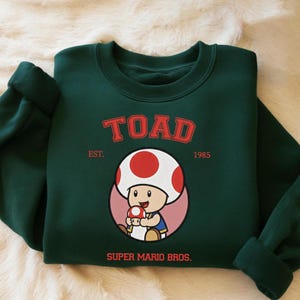 Puede incluir: Sudadera verde oscuro con un diseño de Toad de Super Mario Bros. El diseño incluye la palabra "TOAD" en rojo, el año "1985" y el texto "SUPER MARIO BROS."
