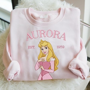 以下が含まれることがあります： ピンク色のスウェットシャツ。ピンク色の刺繍で「AURORA」という名前が描かれ、その下にプリンセスのイラストが描かれています。イラストは、ピンクのドレスを着た金髪のプリンセスです。「EST. 1959」の文字も刺繍されています。